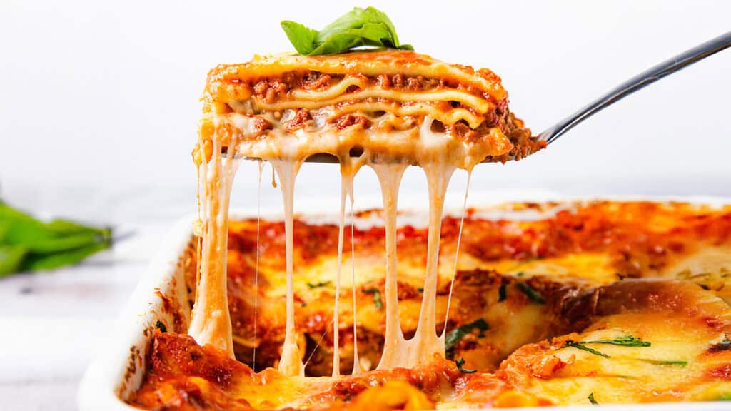 A slice of cheesy lasagna is being lifted from a baking dish, showcasing layers of pasta, rich meat sauce, and gooey melted cheese stretching impressively. A fresh basil leaf garnishes the appetizing slice, making it a star in the world of recipes.