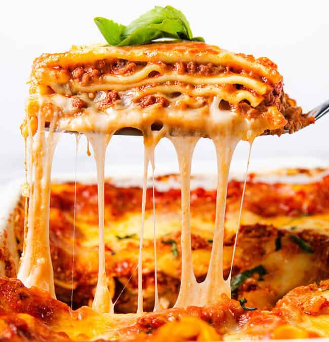 A slice of cheesy lasagna is being lifted from a baking dish, showcasing layers of pasta, rich meat sauce, and gooey melted cheese stretching impressively. A fresh basil leaf garnishes the appetizing slice, making it a star in the world of recipes.