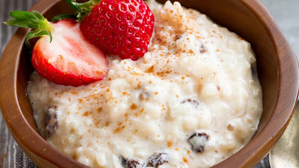 A wooden bowl cradles an old-school treat: creamy rice pudding crowned with a halved strawberry. Cinnamon is dusted over the dessert, revealing plump raisins within. A soft-focus background completes this nostalgic scene.