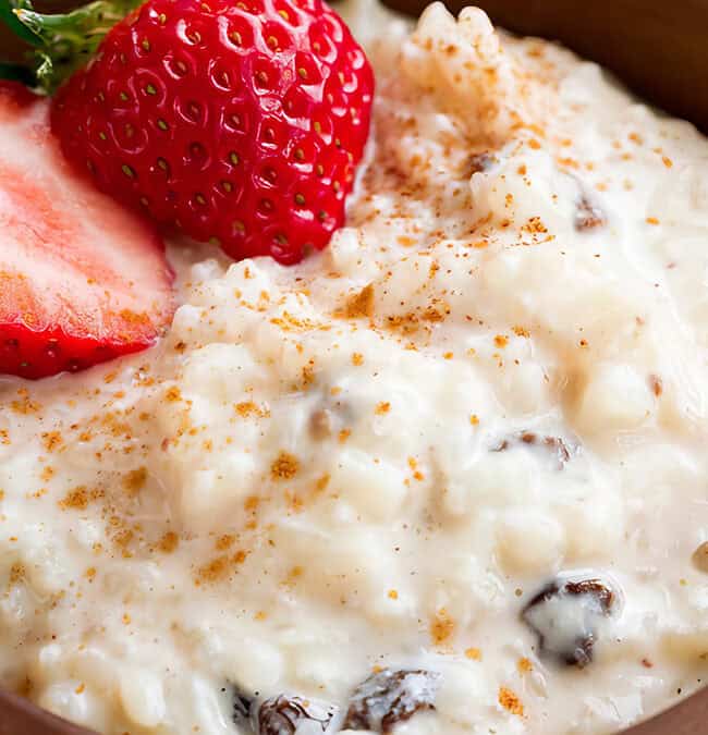 A wooden bowl cradles an old-school treat: creamy rice pudding crowned with a halved strawberry. Cinnamon is dusted over the dessert, revealing plump raisins within. A soft-focus background completes this nostalgic scene.