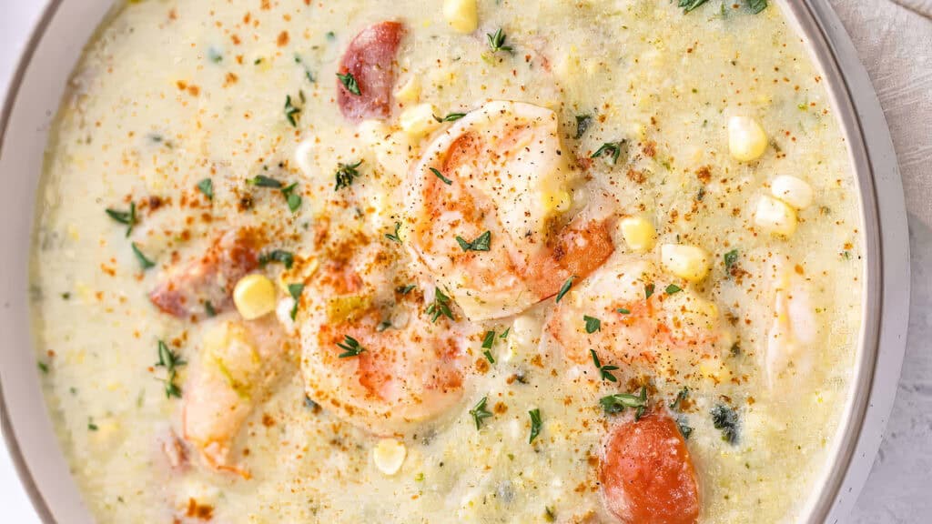 A creamy shrimp chowder, inspired by Southern cuisine, is garnished with fresh herbs and paprika. The dish boasts pieces of shrimp, corn, and potatoes, creating a rich, colorful appearance with a hint of warm spices sprinkled on top.