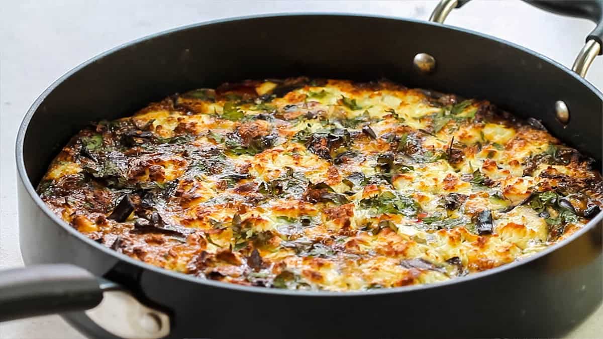 In this enticing frittata recipe, a black skillet houses a golden-brown masterpiece with vibrant vegetables and fresh herbs. The skillet's handle is artfully positioned in the foreground, resting on a light-colored surface—a true testament to skilled cooking.