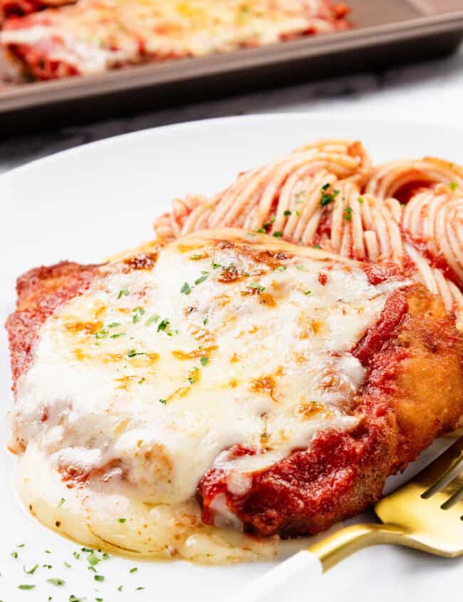 A plate of classic Chicken Parmesan topped with melted cheese and tomato sauce, complemented by spaghetti marinara. Freshly chopped herbs adorn the dish, with a fork resting beside it. Another savory serving awaits in the background on a baking tray.