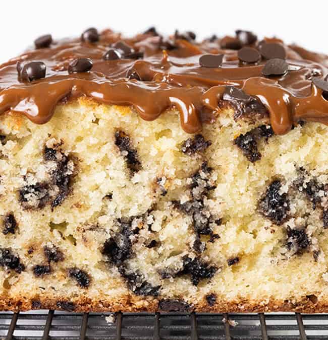 A close-up of a loaf cake with chocolate chips, topped with glossy caramel and sprinkled with more chocolate chips. It's resting on a wire cooling rack. The cake appears moist and rich with a golden-brown crust.