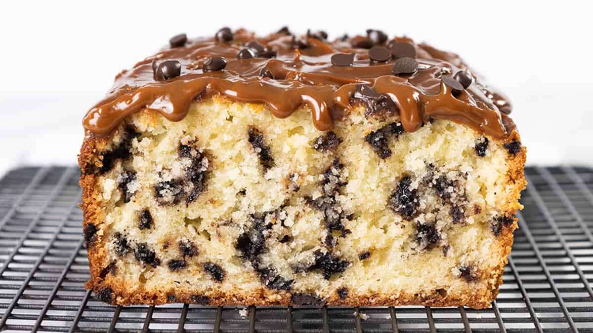 A close-up of a loaf cake with chocolate chips, topped with glossy caramel and sprinkled with more chocolate chips. It's resting on a wire cooling rack. The cake appears moist and rich with a golden-brown crust.