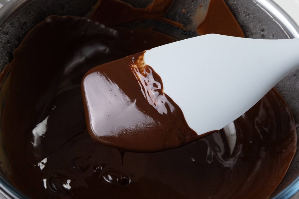 A spatula stirring melted dark chocolate in a bowl creates the perfect base for mousse. The chocolate appears smooth and glossy, coating the spatula as it's being mixed.