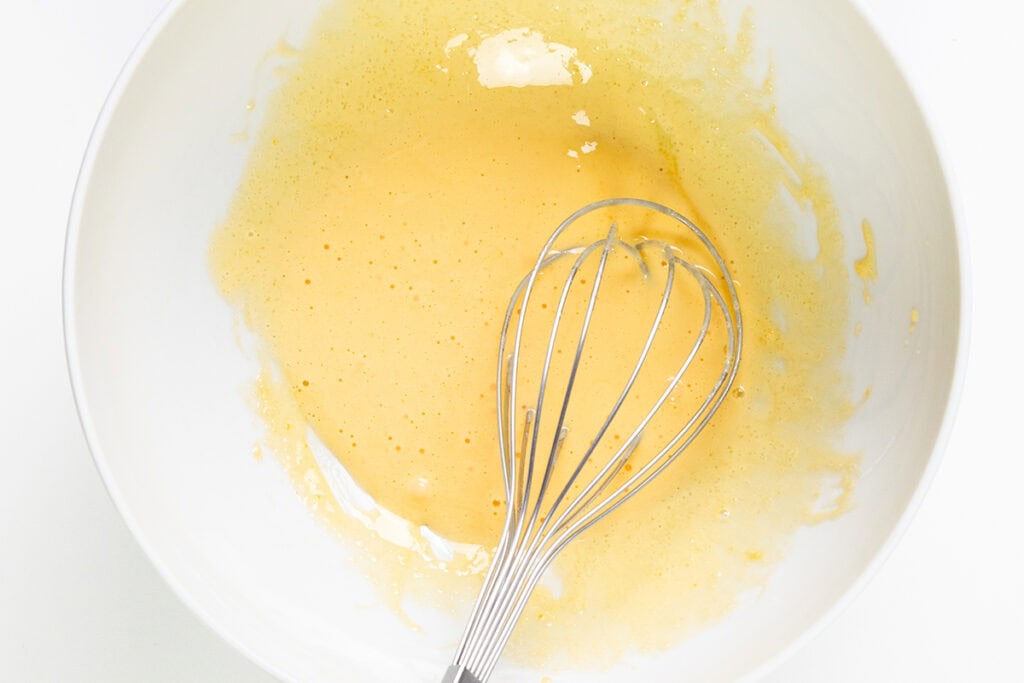 A metal whisk rests in a white bowl containing a yellow, frothy, beaten egg mixture, ready for an indulgent dessert like chocolate mousse. The bowl's interior is partially coated with splash marks, all set against a soft white surface perfect for crafting your next recipe masterpiece.