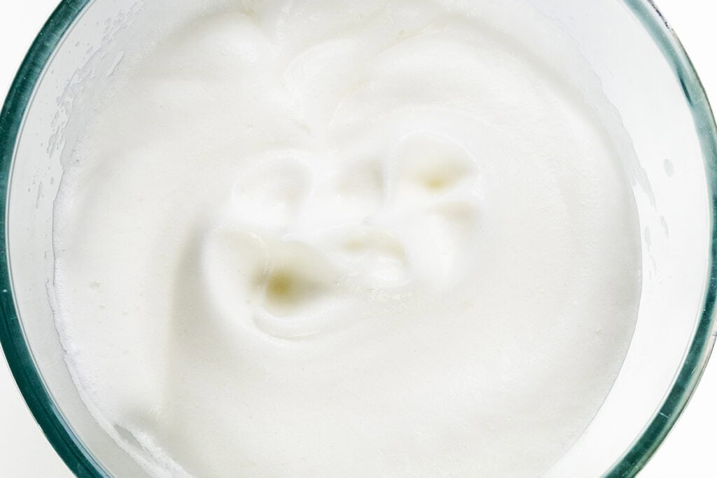 A glass bowl filled with fluffy, whipped egg whites resembles a rich chocolate mousse. The mixture is glossy and smooth, with slightly stiff peaks visible on the surface. The white background emphasizes the creamy texture.