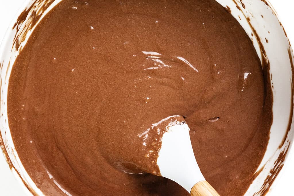 A mixing bowl brims with smooth, dark chocolate cake batter, resembling the richness of mousse. A white spatula with a wooden handle rests within, partially submerged. The bowl's edges are slightly smeared with batter.