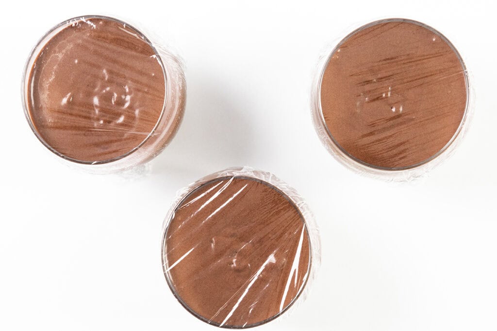 Three glasses of rich chocolate mousse, viewed from above, are elegantly covered with plastic wrap against a pristine white background.