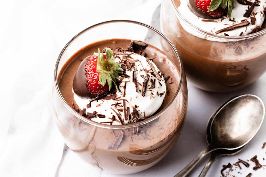 A glass brimming with chocolate mousse is adorned with whipped cream, delicate chocolate shavings, and crowned by a chocolate-covered strawberry. Another glass and two spoons rest in the background on a pristine white surface, inviting indulgence.