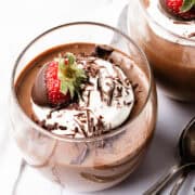 A glass brimming with chocolate mousse is adorned with whipped cream, delicate chocolate shavings, and crowned by a chocolate-covered strawberry. Another glass and two spoons rest in the background on a pristine white surface, inviting indulgence.