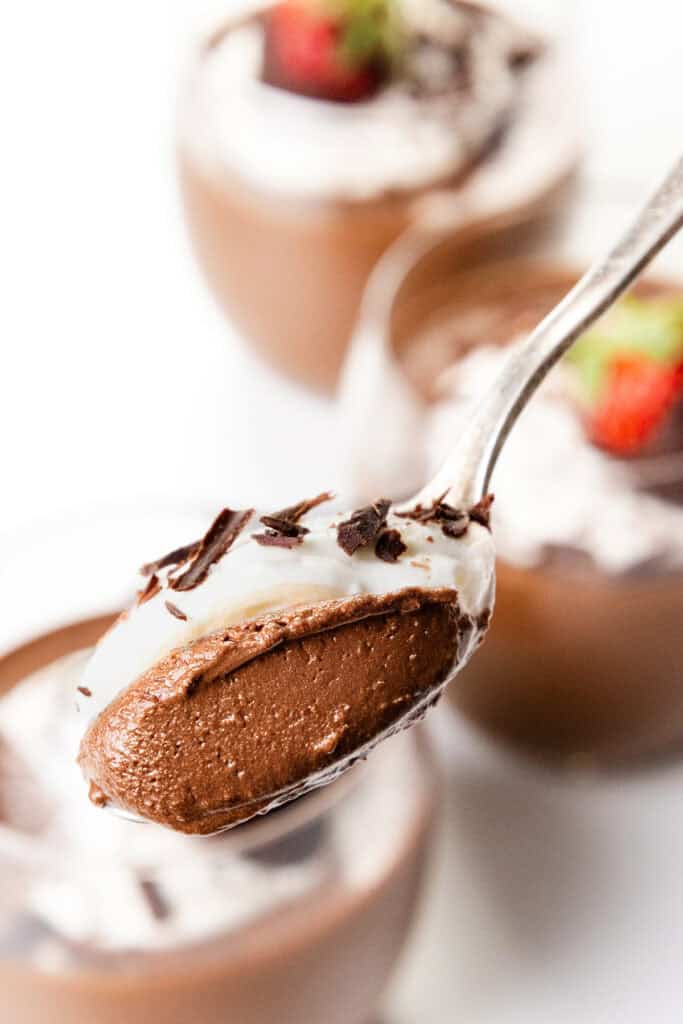 Close-up of a spoon holding silky chocolate mousse topped with whipped cream and delicate chocolate shavings. In the background, blurred glass ramekins are filled with sumptuous mousse, garnished with fluffy whipped cream and fresh strawberries.