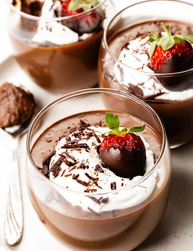 Three elegant glass cups display luscious chocolate mousse crowned with whipped cream, delicate chocolate shavings, and a chocolate-dipped strawberry. A spoon awaits nearby on the light-colored plate, inviting indulgence in these chocolate delights.