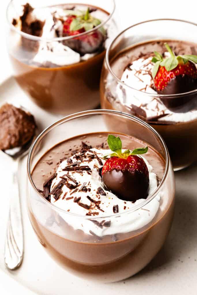 Three elegant glass cups display luscious chocolate mousse crowned with whipped cream, delicate chocolate shavings, and a chocolate-dipped strawberry. A spoon awaits nearby on the light-colored plate, inviting indulgence in these chocolate delights.