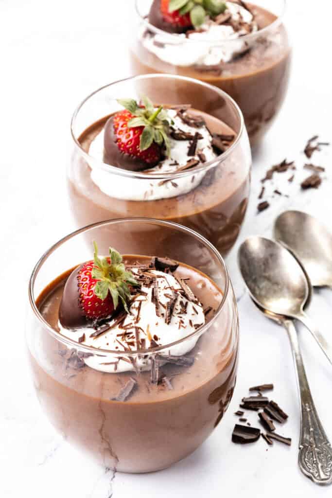 Three glass cups brimming with rich chocolate mousse are topped with airy whipped cream, delicate chocolate shavings, and a strawberry decadently dipped in chocolate. Two elegant silver spoons rest beside the cups on a pristine white marble surface.