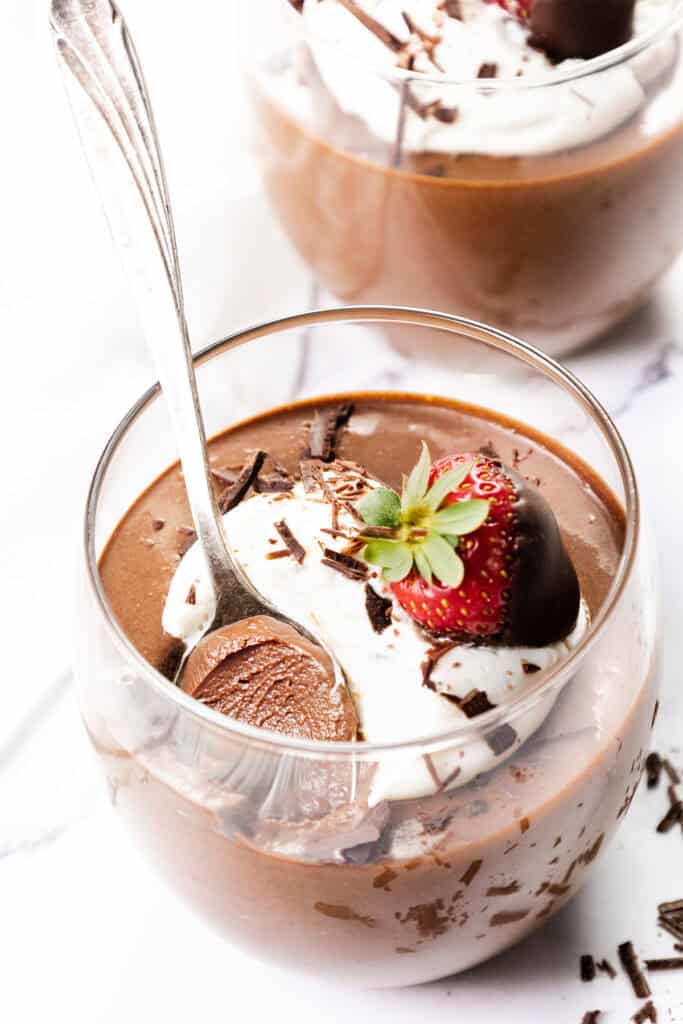 A glass cup brimming with rich chocolate mousse is crowned with whipped cream, delicate chocolate shavings, and a chocolate-dipped strawberry. A spoon playfully peeks from the creamy depths, while another indulgent mousse creation tempts in the background.