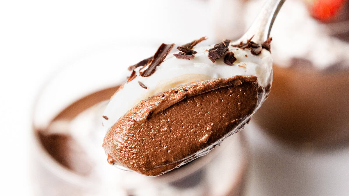 Incredible close-up of a spoonful of chocolate mousse topped with whipped cream and chocolate shavings. A blurred background reveals a glass filled with more mousse, garnished with a strawberry and cream—perfect for sharing with family.