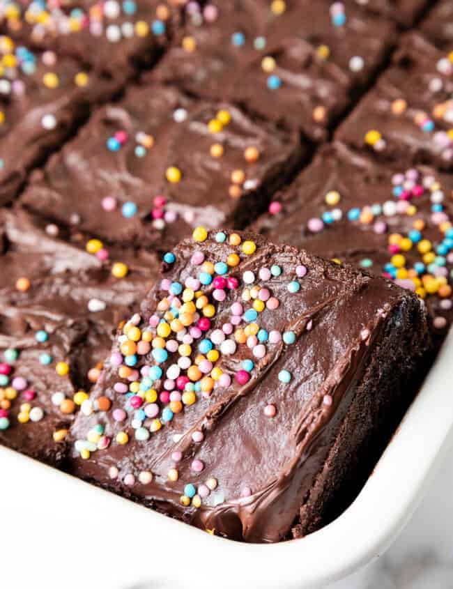 A tray of sliced chocolate brownies topped with colorful sprinkles and featuring a rich, glossy whipped ganache frosting. One piece is slightly raised to reveal the decadent layers. The brownies are displayed in a white ceramic dish reminiscent of a classic chocolate sheet cake.