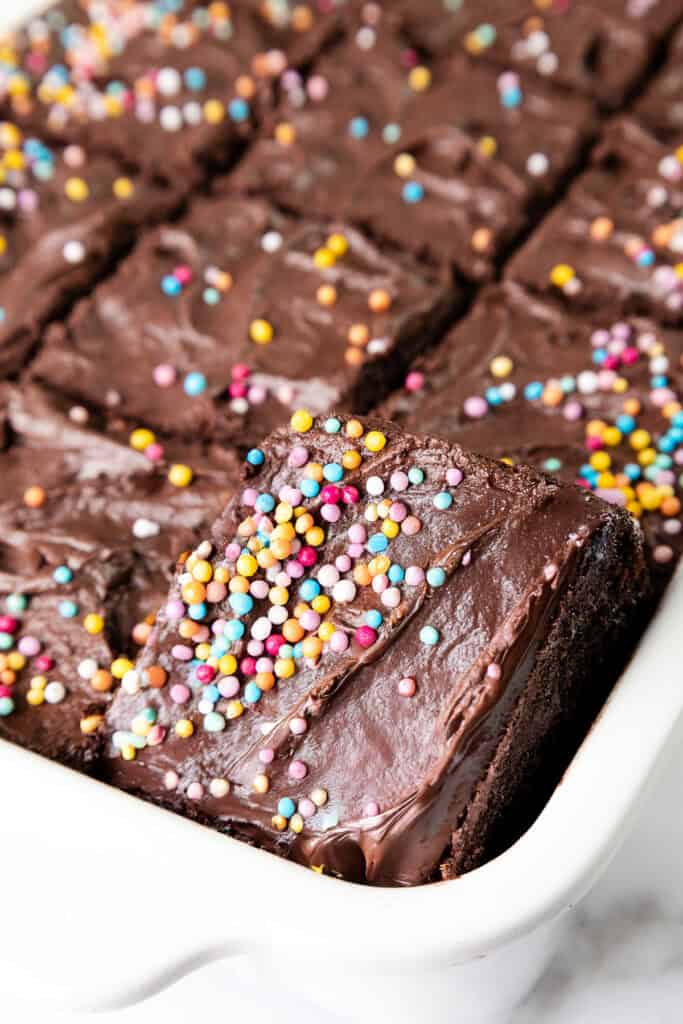 A tray of sliced chocolate brownies topped with colorful sprinkles and featuring a rich, glossy whipped ganache frosting. One piece is slightly raised to reveal the decadent layers. The brownies are displayed in a white ceramic dish reminiscent of a classic chocolate sheet cake.