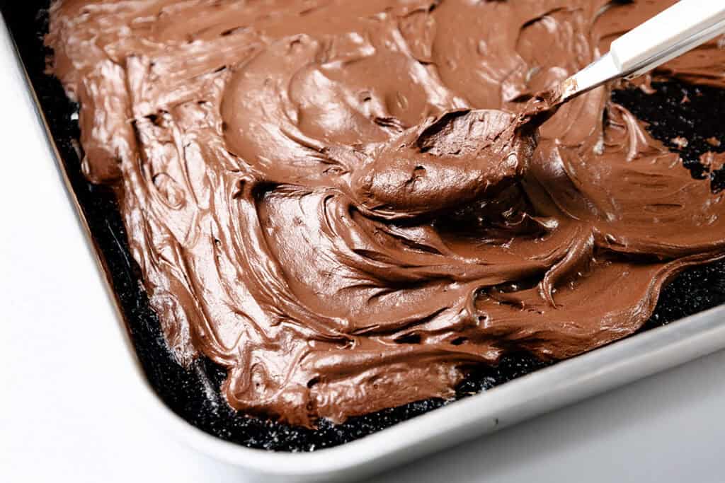 Close-up of a baking pan with whipped ganache frosting being spread over a chocolate sheet cake. A spatula smooths the creamy layer on the surface, making it look delectable on the white countertop.