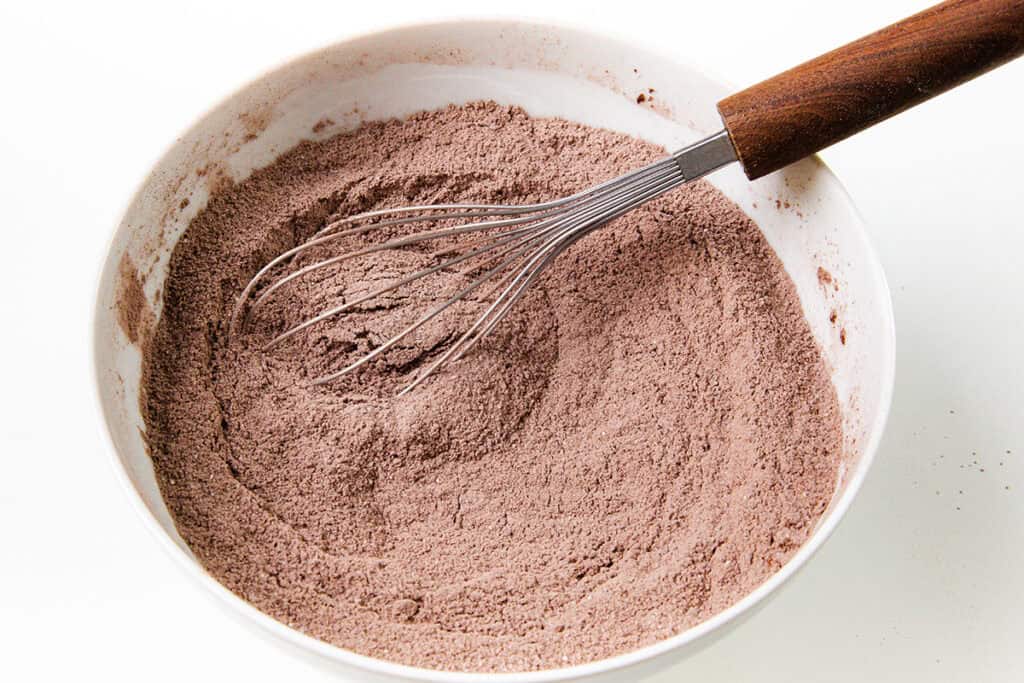 A white bowl filled with chocolate cake mix powder sits invitingly, ready to transform into a delectable chocolate sheet cake. A metal whisk with a wooden handle rests on the mix, eager to whip it into perfection against a plain white backdrop.