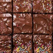 A decadent tray of chocolate sheet cake brownies cut into squares, topped with multicolored sprinkles. The whipped ganache frosting is smooth and glossy, with a rich, creamy texture. Each brownie is evenly decorated with vibrant sprinkles.