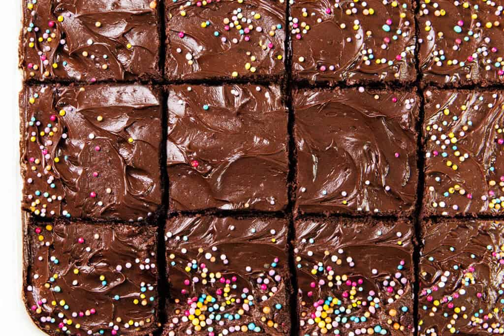 A top view of chocolate frosted brownies cut into squares showcases a decadent chocolate dessert. The smooth whipped ganache frosting, adorned with colorful sprinkles, adds a vibrant touch to the rich, dark surface akin to a tempting chocolate sheet cake.