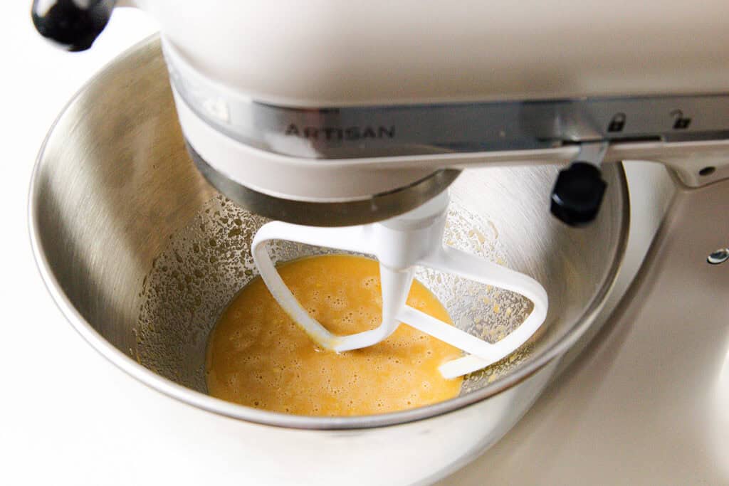 A stand mixer with a paddle attachment is blending a yellow batter in a metal bowl, setting the stage for a delightful Chocolate Sheet Cake. The appliance is primarily white and partially visible from the side.