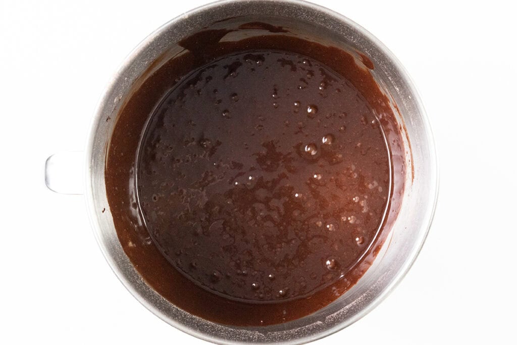 A silver mixing bowl filled with smooth, dark chocolate cake batter is viewed from above. The batter appears glossy and thick, with small bubbles on its surface. The bowl has a handle on the left side.