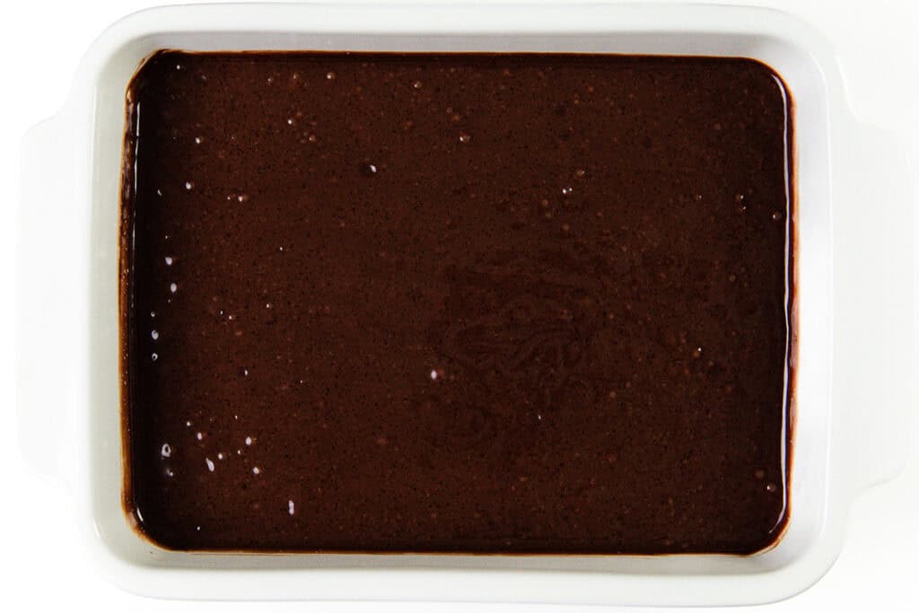 A white rectangular baking dish filled with smooth, dark chocolate cake batter awaits transformation into a decadent Chocolate Sheet Cake. The glossy batter is evenly spread, promising a delightful bake that begs for rich Whipped Ganache Frosting as its crowning glory.