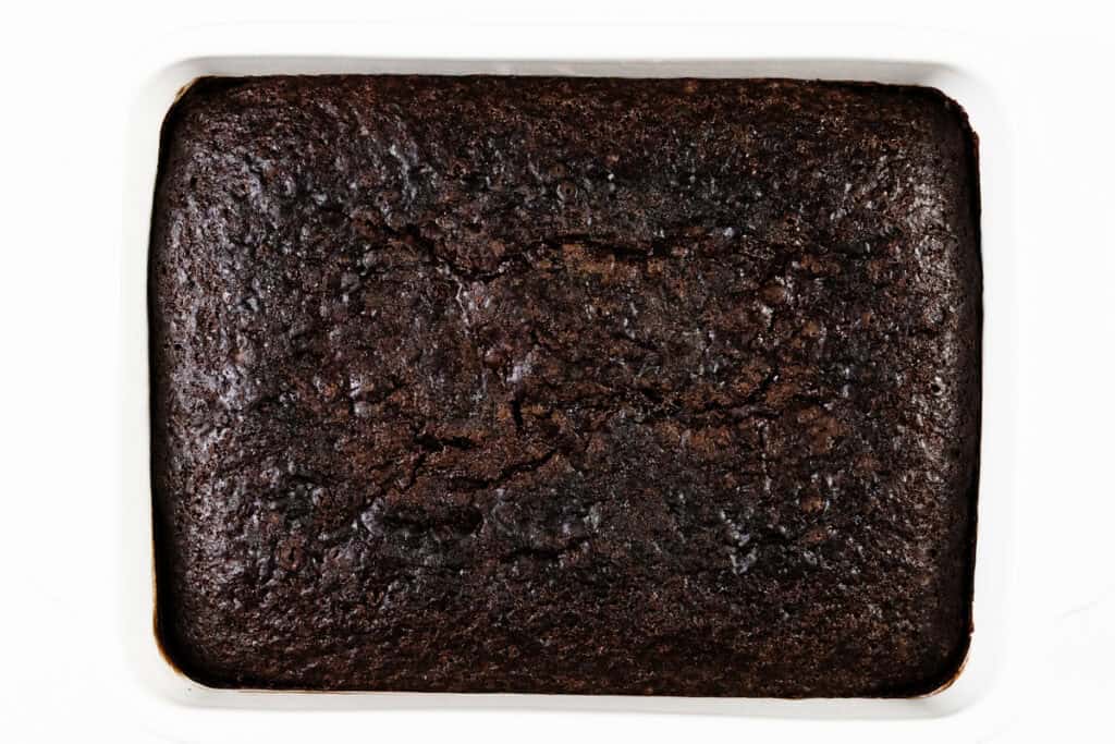 A freshly baked dark chocolate sheet cake sits in a rectangular white baking dish, its surface slightly cracked.