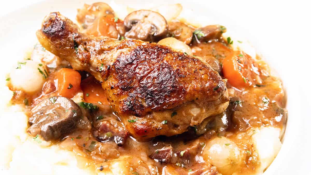 Close-up of a plate featuring a crispy roasted chicken drumstick on a bed of thick stew with carrots, mushrooms, and onions, garnished with herbs. This family favorite is served over creamy mashed potatoes—one of those incredible recipes you'll want to make again.