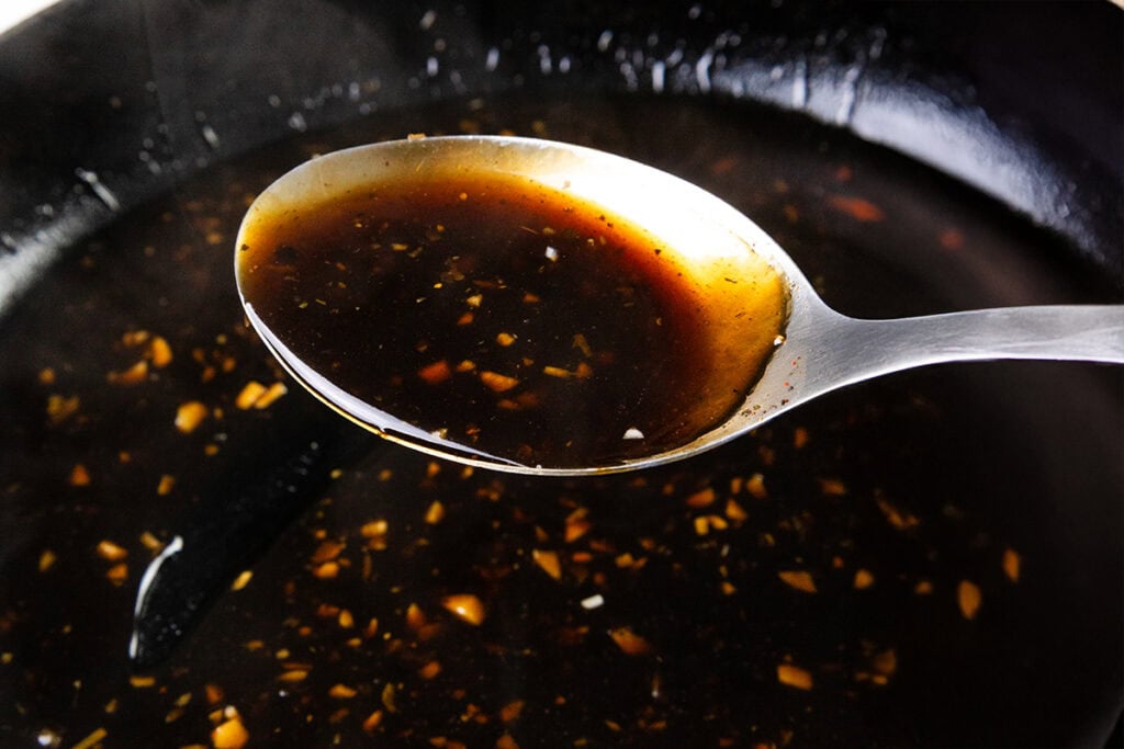 Close-up of a metal spoon lifting a Bourbon Glaze from a black pan. The sauce, perfect for pairing with crispy skin chicken, contains visible minced garlic and small spices, creating a textured look. The background is softly blurred, highlighting the spoon and sauce.