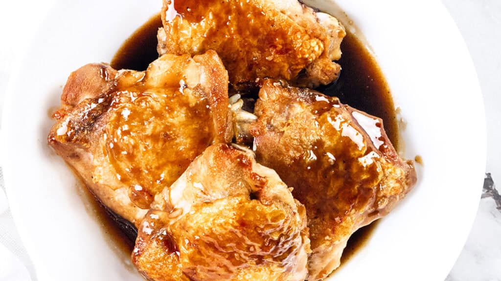 Four pieces of glazed, crispy chicken in a white bowl, coated with a shiny, brown sauce.