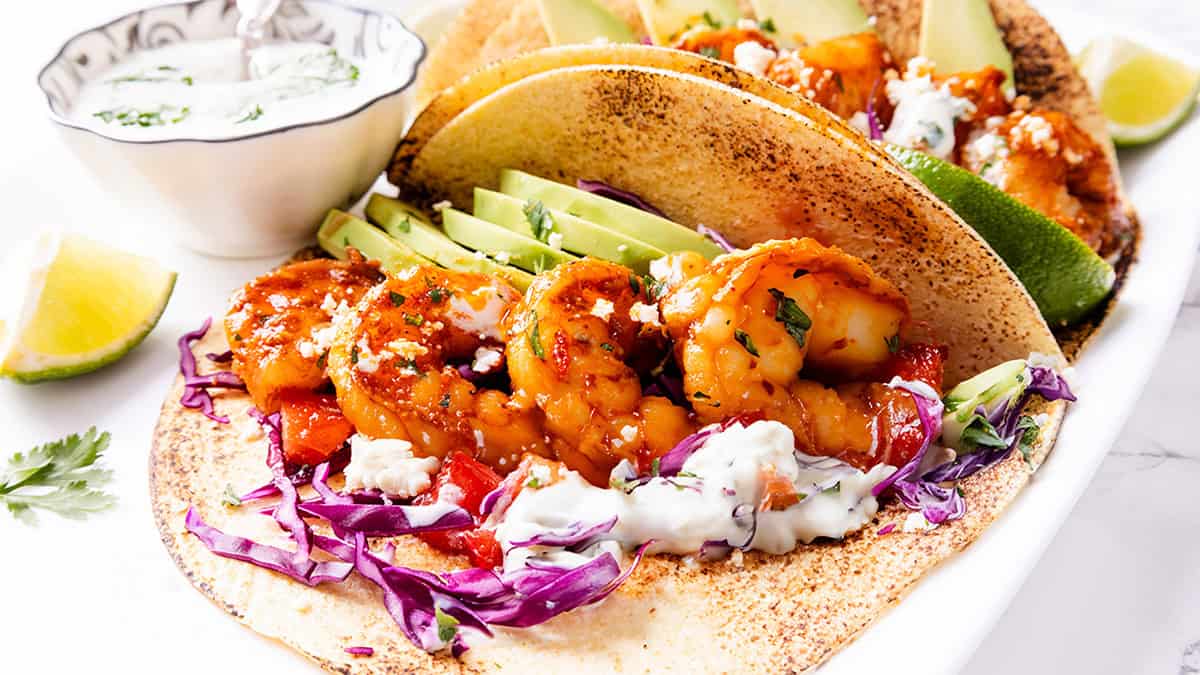 A close-up of shrimp tacos filled with grilled shrimp, sliced avocado, shredded purple cabbage, and drizzled white sauce. The tacos are on a tortilla, garnished with cilantro, with a bowl of sauce and lime wedges in the background.