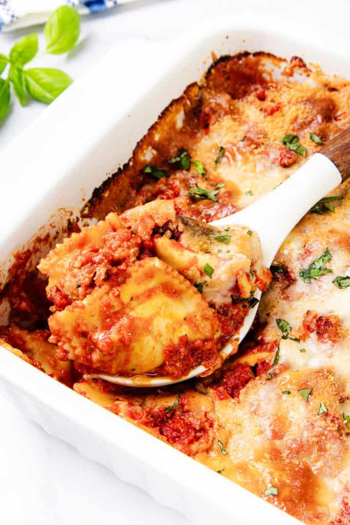 A serving of ravioli casserole is being scooped from a white baking dish. Topped with melted cheese and tomato sauce, it's garnished with fresh basil leaves. The golden-brown edges hint at its freshly baked perfection.