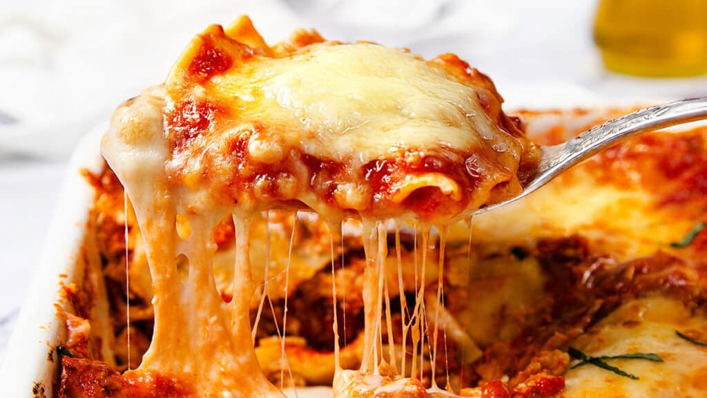 Close-up of a slice of Italian Sausage Ravioli Casserole being lifted from a dish. This classic Italian recipe boasts a rich layer of melted cheese stretching with mozzarella strands. The dish features a tomato-based sauce and hints of fresh basil on top.