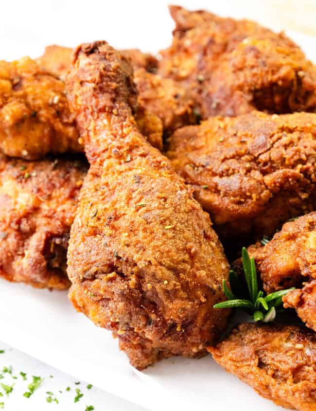 A close-up of KFC-style crispy fried chicken pieces on a white surface, garnished with fresh rosemary. The golden-brown skin is seasoned and textured, suggesting a flavorful and crunchy coating.