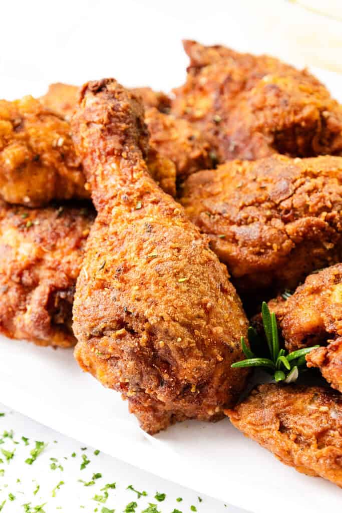 A close-up of KFC-style crispy fried chicken pieces on a white surface, garnished with fresh rosemary. The golden-brown skin is seasoned and textured, suggesting a flavorful and crunchy coating.