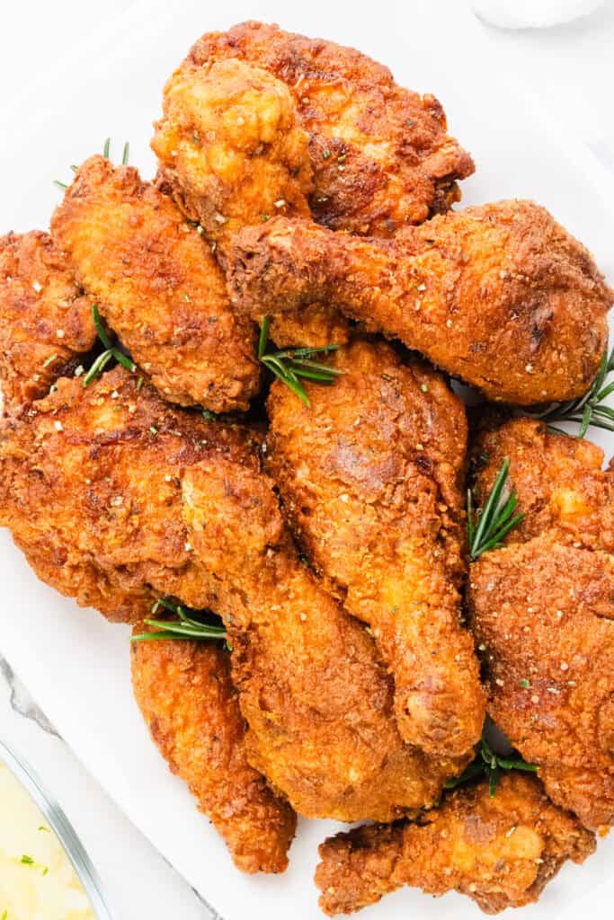 A plate of KFC-style, golden-brown fried chicken drumsticks and wings garnished with fresh rosemary, resting on a white surface.