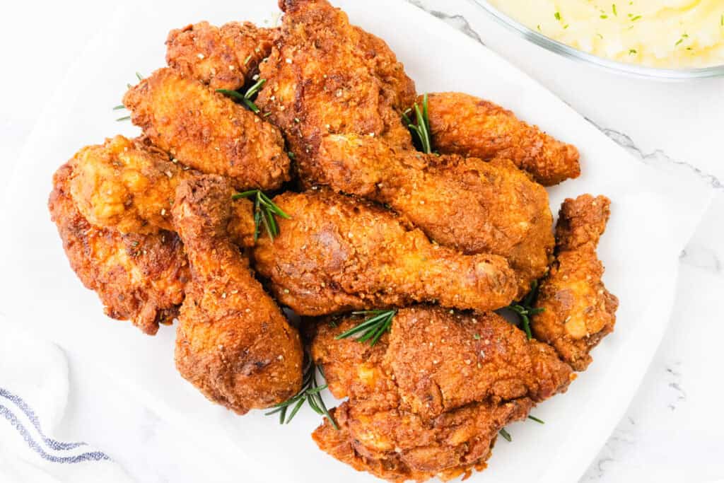 A plate of golden-brown, KFC-style fried chicken pieces garnished with rosemary sprigs is displayed on a white surface. A bowl of creamy mashed potatoes is partially visible in the background.