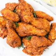 A plate of golden-brown, KFC-style fried chicken pieces garnished with rosemary sprigs is displayed on a white surface. A bowl of creamy mashed potatoes is partially visible in the background.