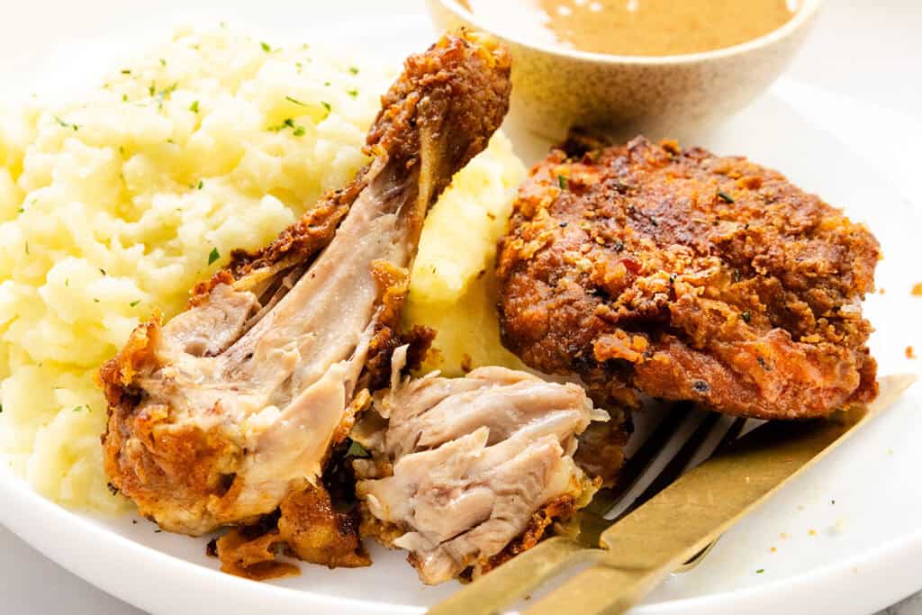 A white plate holds mashed potatoes garnished with herbs, a serving of KFC-style fried chicken, and a partially eaten drumstick. A fork rests nearby, while a small bowl of gravy lingers in the background, ready to enhance the savory experience.