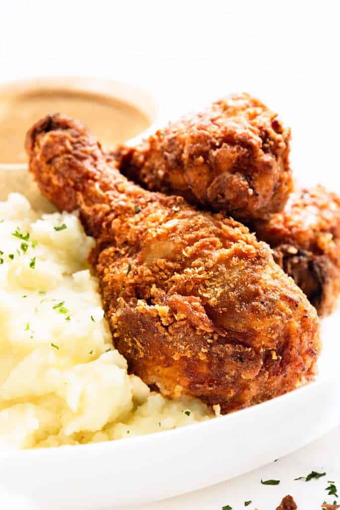 Plate with crispy KFC Fried Chicken Recipe drumsticks, creamy mashed potatoes sprinkled with herbs, and a side of savory gravy in a small dish. Served on a pristine white plate.