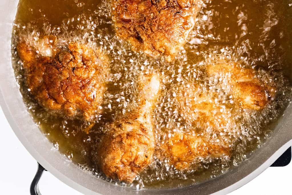 Golden brown pieces of KFC-style fried chicken sizzle in hot oil, creating bubbles and a crisp texture. The close-up image captures the sizzling action of the cooking process in a pan, highlighting every golden crunch.