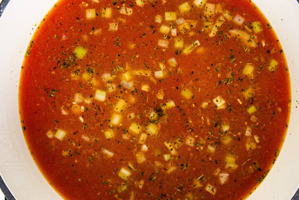 A close-up of tomato soup with visible chunks of onion, herbs, and tender pasta. The soup, reminiscent of a classic Veneta dish, has a reddish hue with ingredients elegantly distributed throughout the liquid, creating a textured and appetizing appearance.