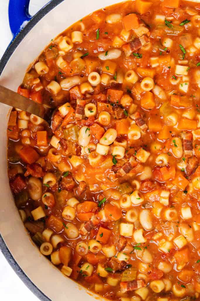 A pot of hearty Italian soup reminiscent of filled with ditalini pasta, beans, diced tomatoes, carrots, celery, and herbs. A ladle dips into the thick, colorful mixture, showcasing a delightful variety of vegetables and textures.