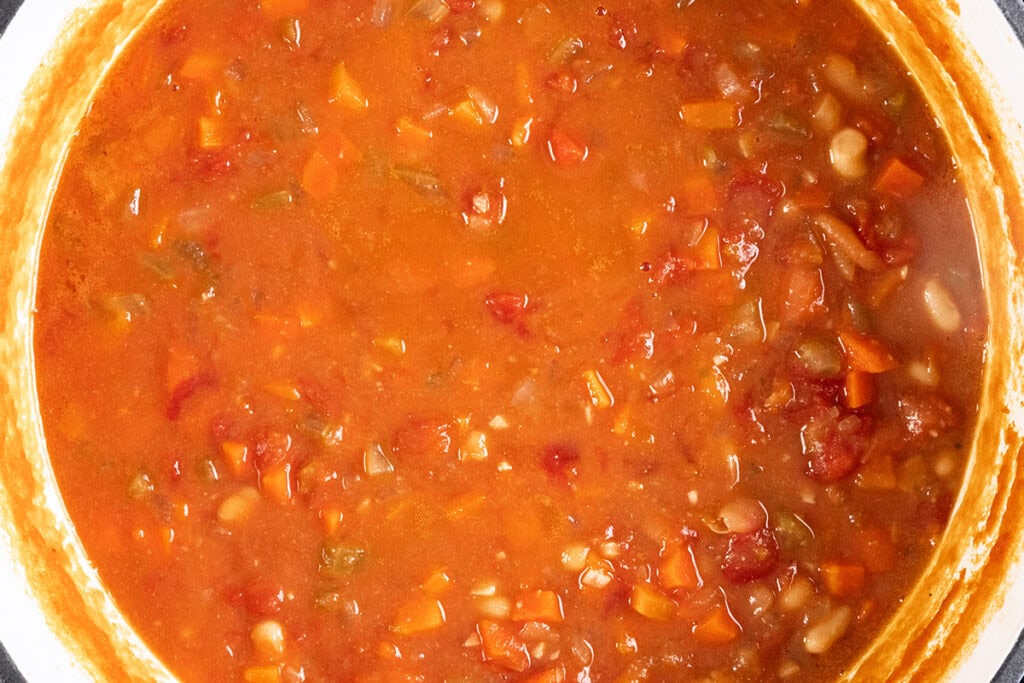 A close-up of a pot filled with vibrant vegetable soup reminiscent of Pasta Fagioli alla Veneta. The soup features chunks of tomatoes, carrots, beans, and other vegetables in a rich red broth. The pot's rim is partially visible.