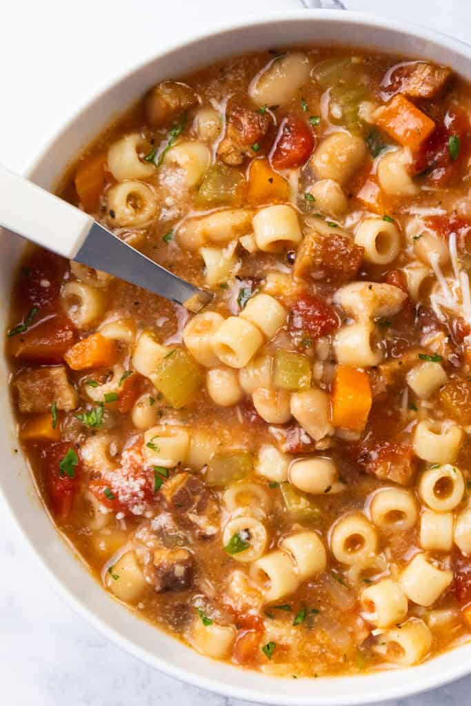 A bowl of pasta soup featuring ditalini pasta, diced tomatoes, carrots, celery, and herbs. A spoon rests in the bowl, revealing a hearty and colorful mix. The slightly reddish broth appears delectably thick.